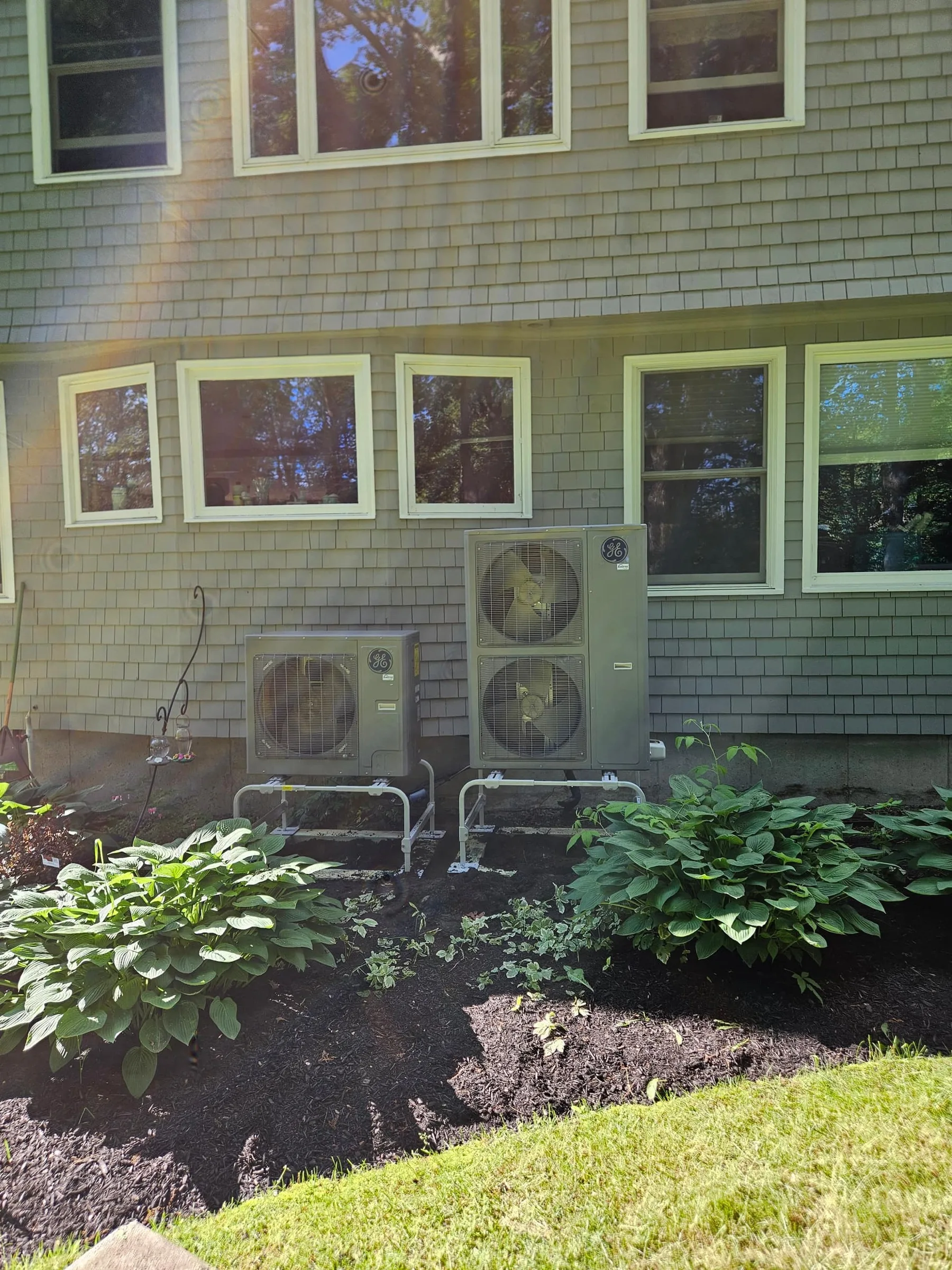 Two GE outdoor heat pumps stand on brackets in front of a shingled house.